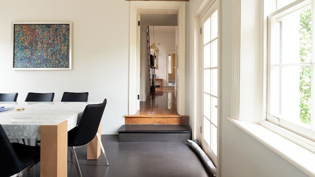 Un espace repas moderne avec une table en marbre et des chaises noires, une grande peinture abstraite au mur et une vue sur un couloir menant à un escalier.
