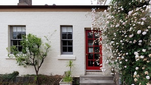 Un bâtiment blanc avec une porte et des fenêtres rouges, un petit arbre et un jardinier avec des plantes.