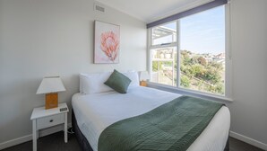 A hotel room with a large bed, a nightstand with a lamp, a green throw blanket, and a window with a view of the city. - Sea You Soon: Views, Parking & WiFi (Montello)