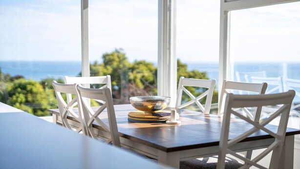 A dining table with white chairs and a decorative bowl, set against a backdrop of a scenic view through large windows. - Sea You Soon: Views, Parking & WiFi (Montello)