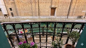 Un balcón con barandilla de hierro forjado, plantas en macetas y vista a un edificio de ladrillo con una puerta.