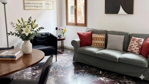 Una sala moderna con un sofá gris, una mesa redonda de madera, un jarrón con flores, una fotografía enmarco de un paisaje urbano y un cuadro abstracto grande.