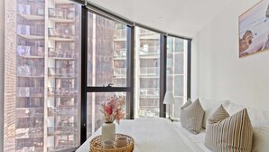 Un dormitorio moderno con ventanas amplias, una cama con almohadas a rayas, un florero con flores y una bandeja de mimbre con tazas.