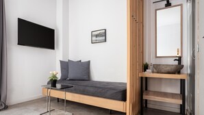 A modern, minimalist room with a bed, a mounted TV, a small table with a plant, a wooden shelf with a mirror, and a picture on the wall. - Acropolis Cozy Living (Athina)