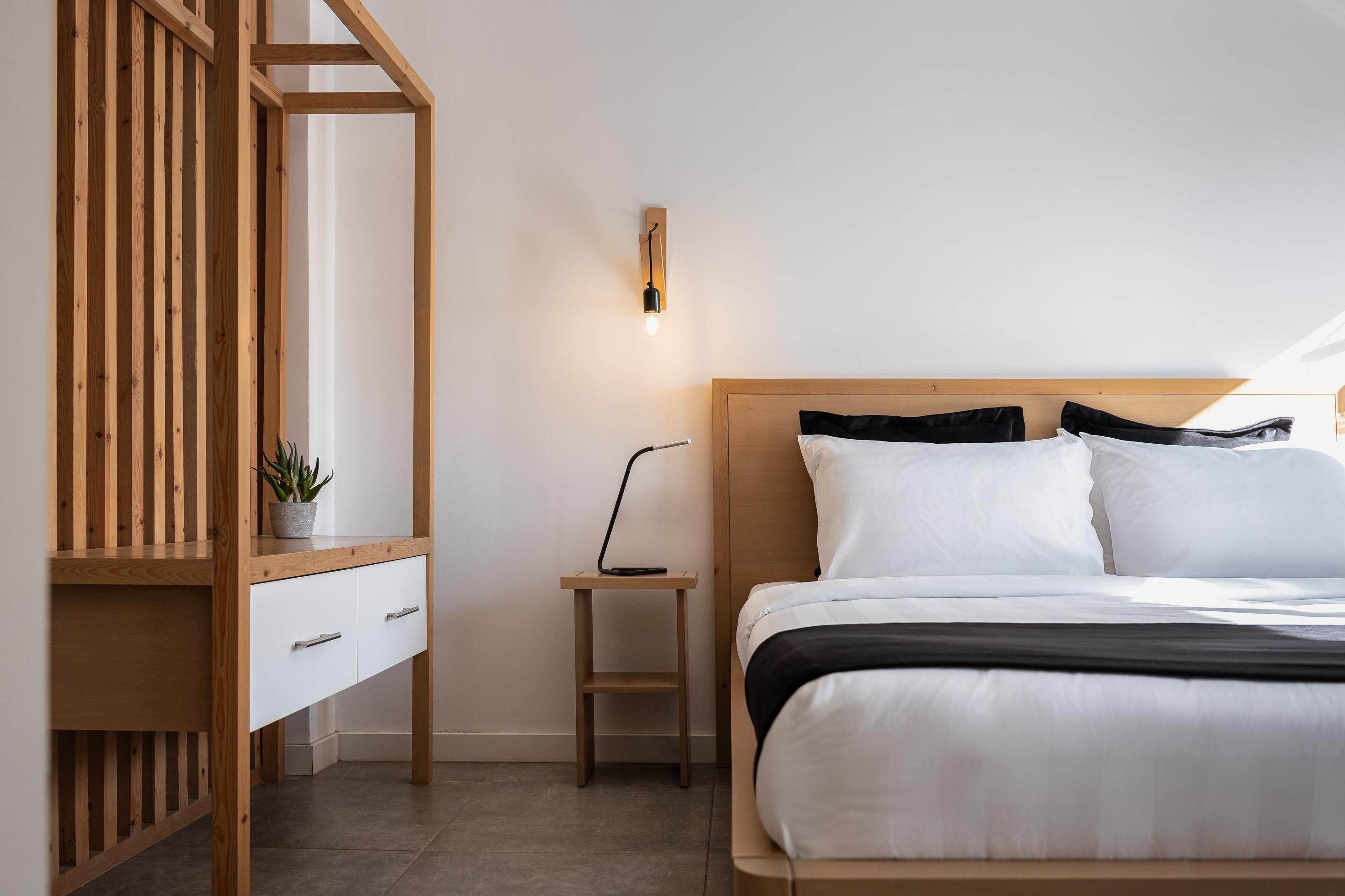 A bedroom with a wooden bed frame, white bedding, a bedside table with a lamp, and a wooden wardrobe with a plant.
