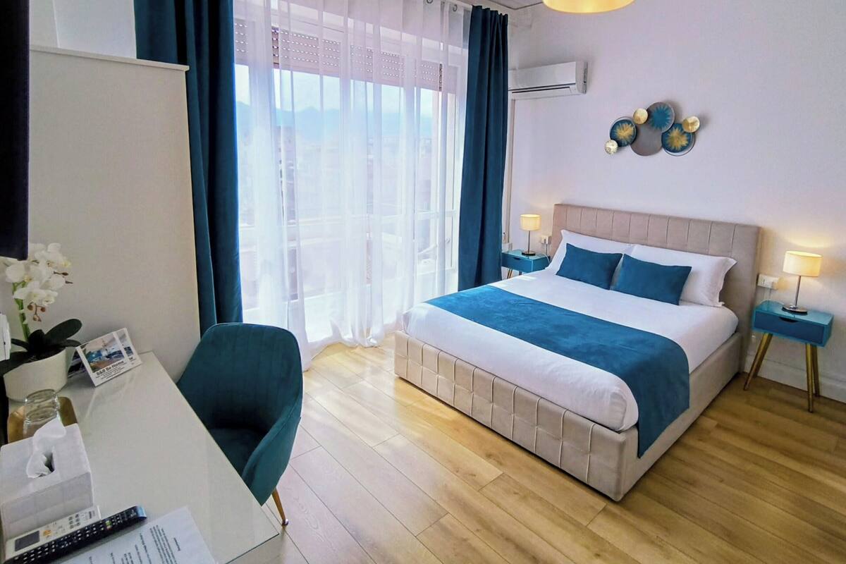 A hotel room with a bed, a desk, a chair, a window with curtains, and a wall-mounted air conditioner.