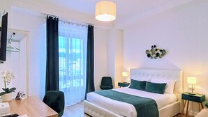 Et hotelværelse med seng, sengeborde, et skrivebord, en stol, en potteplante og et vindue med gardiner.