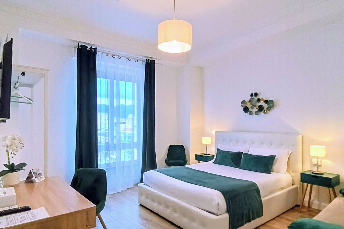A hotel room with a bed, bedside tables, a desk, a chair, a potted plant, and a window with curtains.