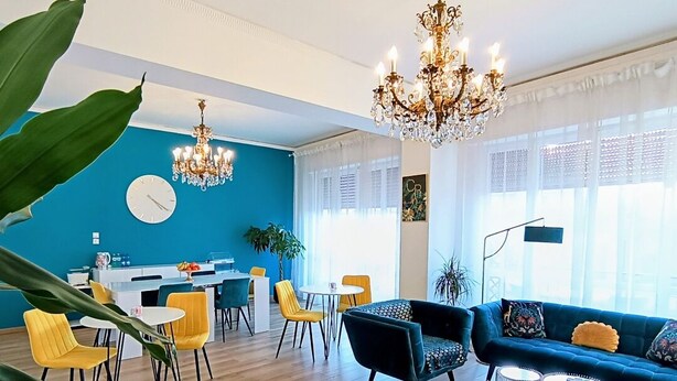 En moderne stue med en blå accentvæg, en lysekrone, en sofa og et spiseområde med gule stole.