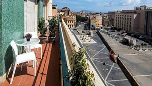 En balkon med et bord, stole, blomsterkrukker og udsigt til bybillede med bygninger og parkeringsplads.