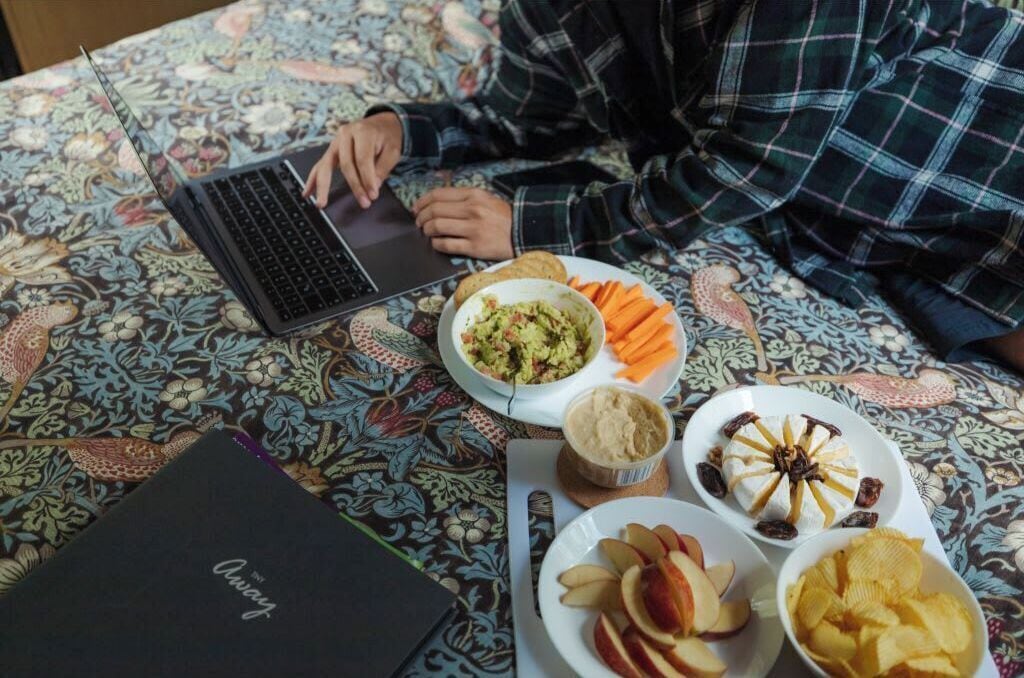En person sitter vid ett bord och använder en bärbar dator. På bordet ligger en mönstrad duk och det finns mat, bland annat en skål med dippsås, äppelklyftor och chips.