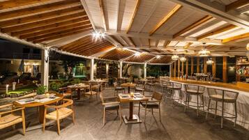 An outdoor dining area with wooden tables and chairs, a bar, and a covered seating area with a view of the garden.