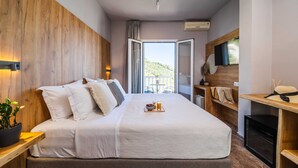 A modern hotel room with a large bed, a wooden headboard, a TV, a small table with a plant, and a balcony with a view. - Skiathos Avaton Garden (Skiathos)