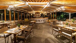 An outdoor dining area with wooden tables and chairs, a bar in the center, and large windows. - Skiathos Avaton Garden (Skiathos)
