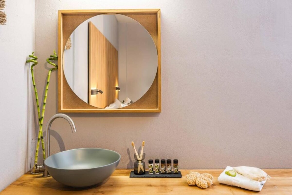 A bathroom with a round mirror, a bowl sink, bamboo plants, and a shelf with toiletries.