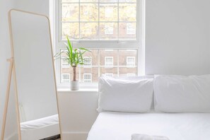 Een minimalistische slaapkamer met een bed, witte kussens, een staand spiegel en een plant in een pot bij een raam.