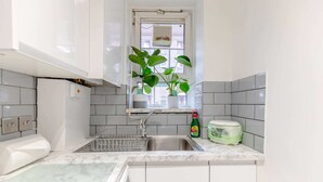 A kitchen with white cabinets, a marble countertop, a window with plants, a sink, and a dish soap bottle. - Central & Stylish 1BD Flat - 8mins to Greenwich! (London)