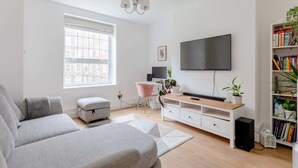 A modern living room with a grey sofa, a pink chair, a white desk, a TV, and a bookshelf. - Central & Stylish 1BD Flat - 8mins to Greenwich! (London)