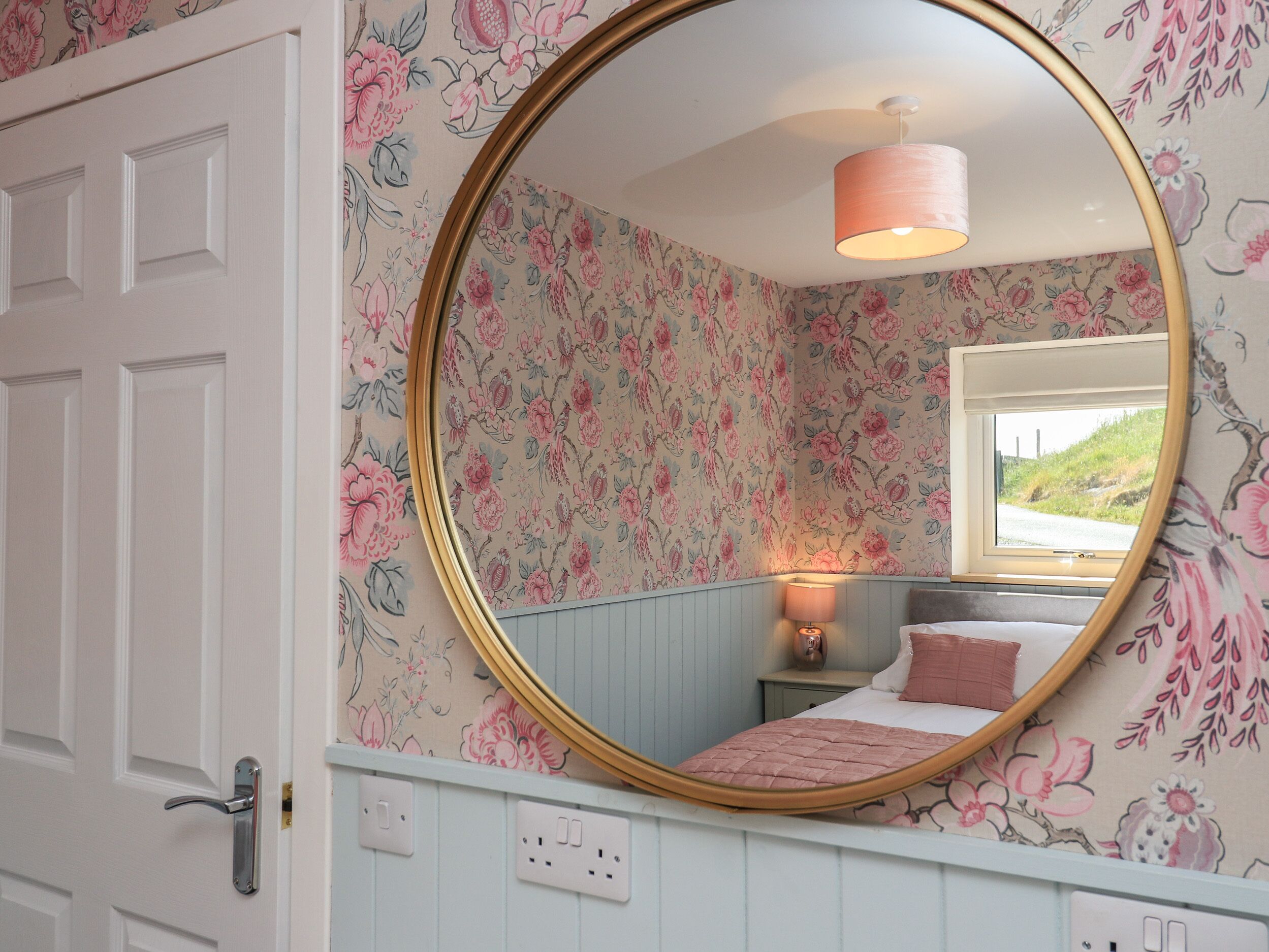 A bedroom with floral wallpaper, a round mirror, a bed with a pink quilt, a nightstand with a lamp, and a window with a view of greenery.