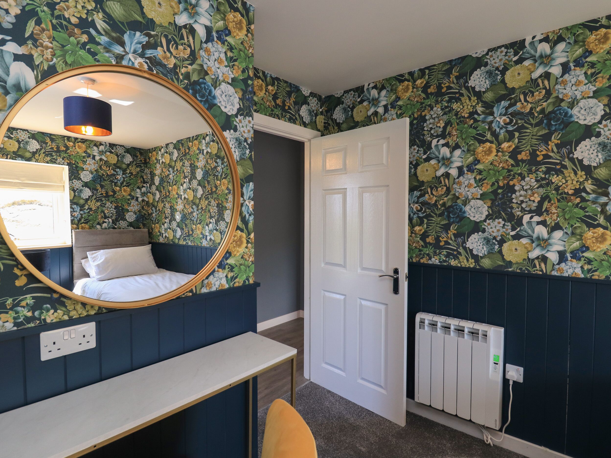 A bedroom with a bed, a round mirror, a bedside table, a radiator, and floral wallpaper.