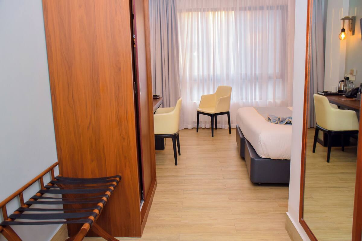 A hotel room with a wooden staircase, a bed, a desk, two chairs, and a large window with sheer curtains.
