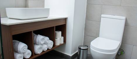 A bathroom with a white sink, wooden vanity, towel storage, toilet, and trash can.