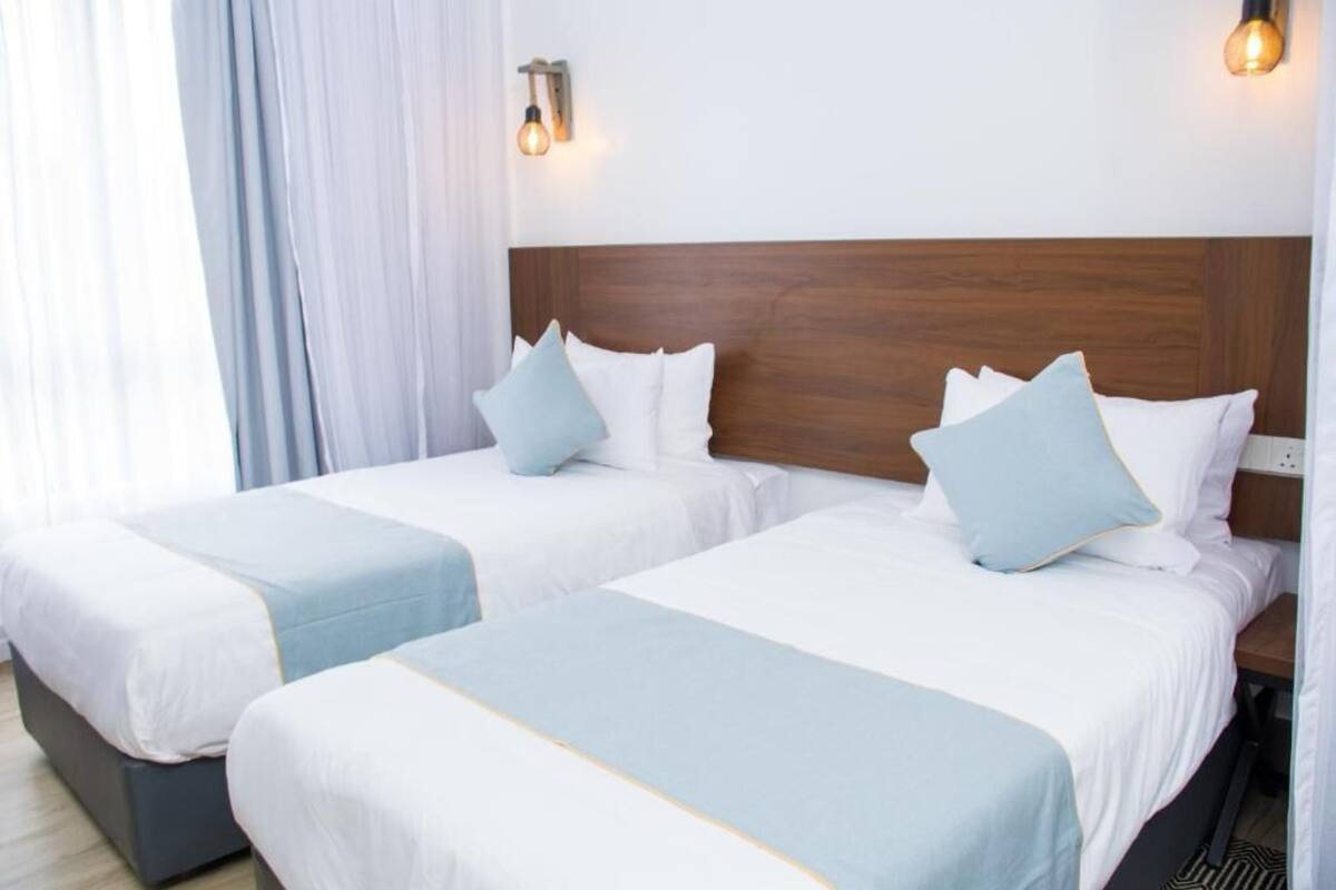 A hotel room with two beds, each with white bedding and light blue accents. There are wall-mounted light fixtures on either side of the headboard.