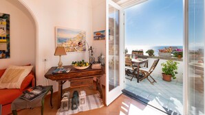 A cozy living room with a wooden desk, a comfortable red sofa, and a view of the terrace with potted plants and a table. - Casa Peppenella in Positano (Positano)