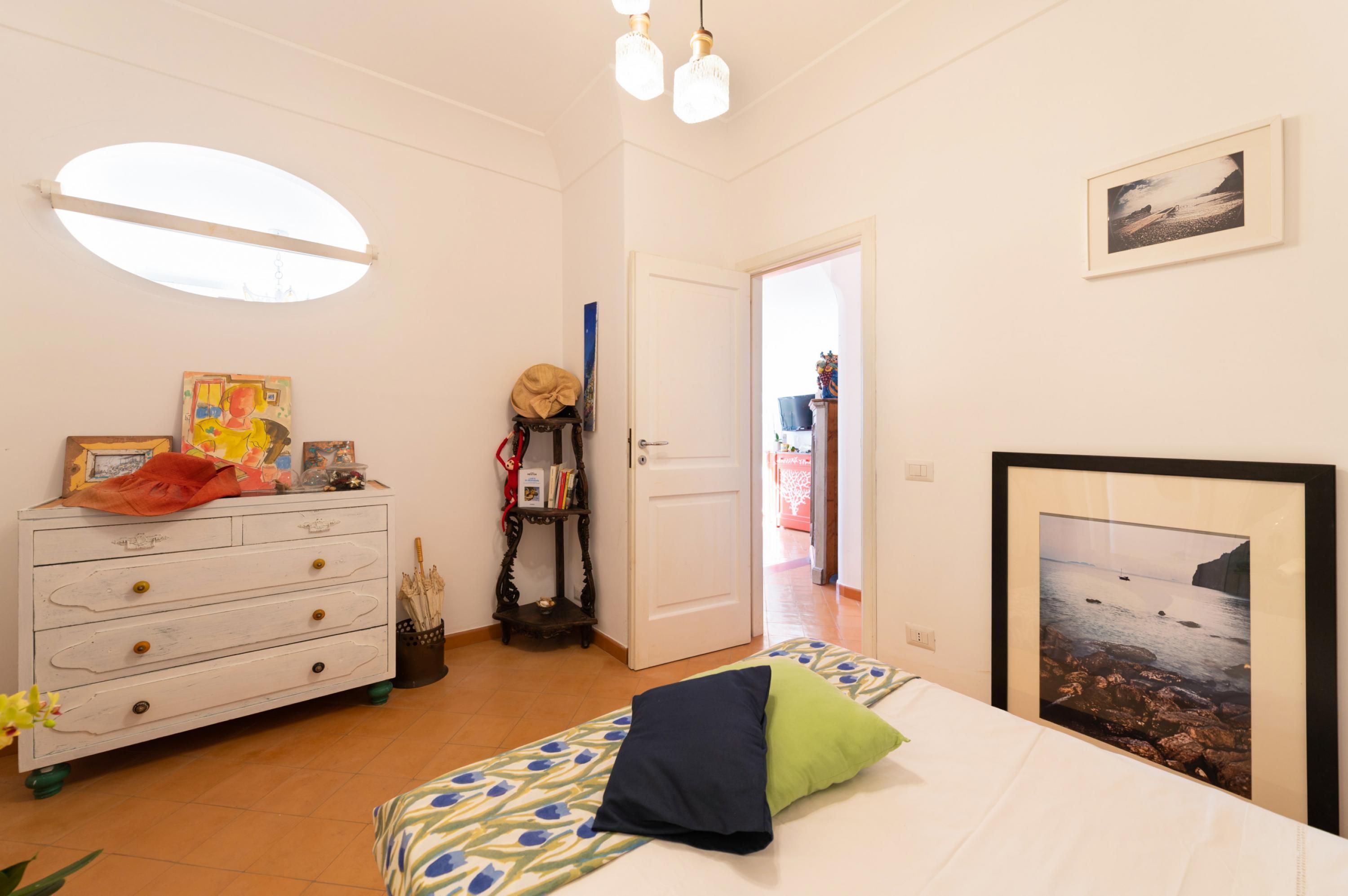 Un dormitorio con cama, cómoda, cómoda con espejo, un cuadro enmarcado en la pared y una puerta que da a otra habitación.