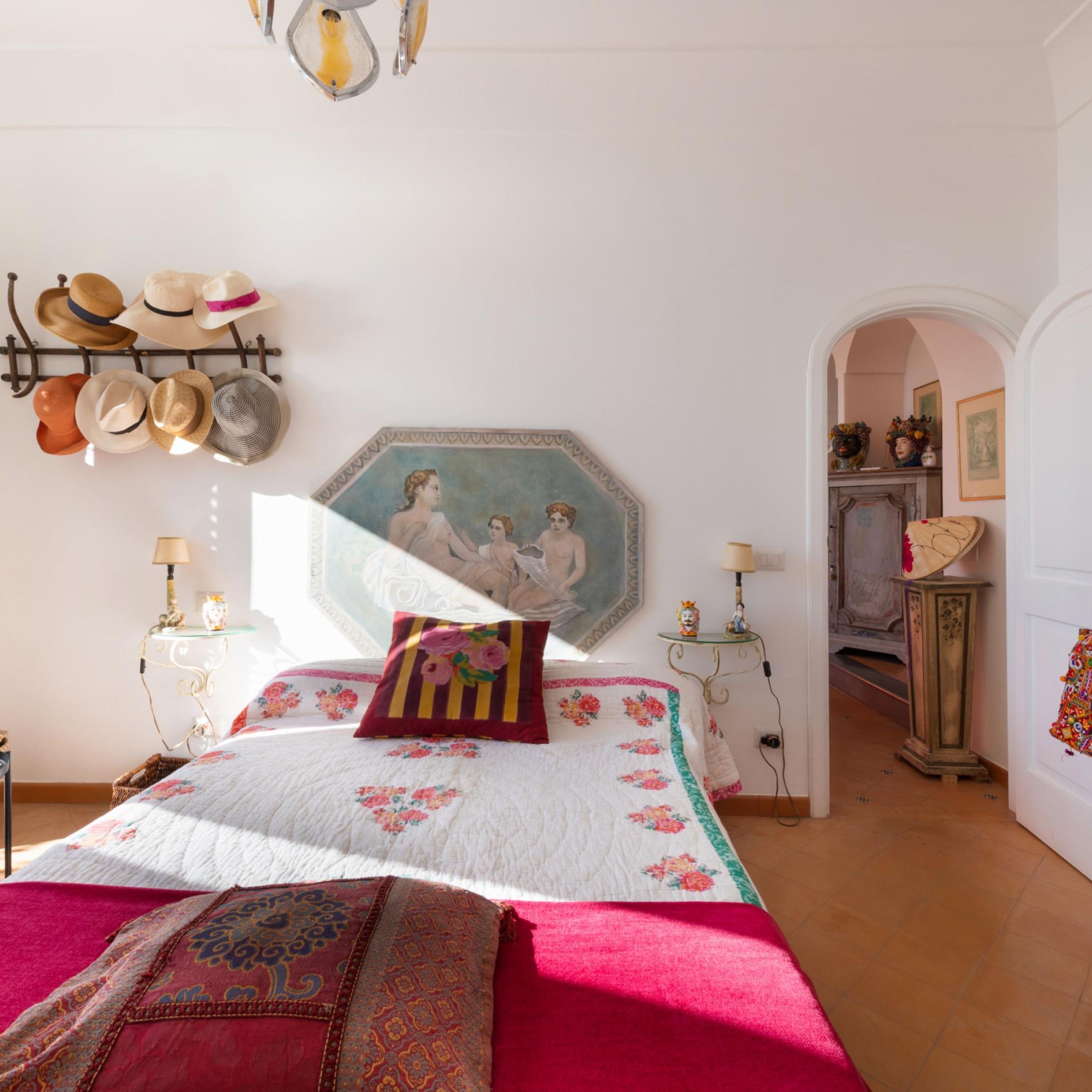 A bedroom with a bed, a wall-mounted hat rack, a painting, and a hallway leading to another room.