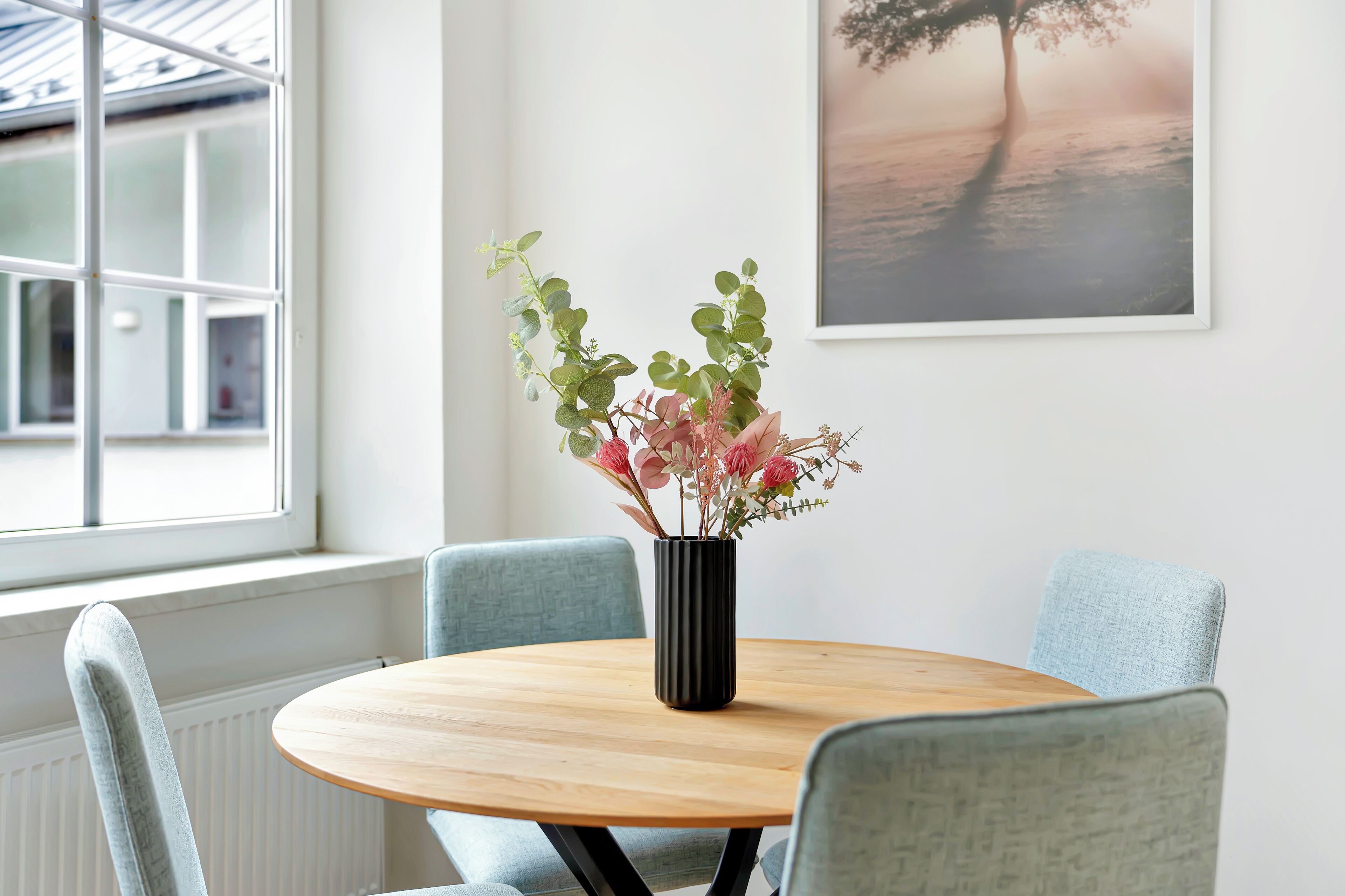 A round wooden table with a black vase containing flowers, surrounded by light blue upholstered chairs.