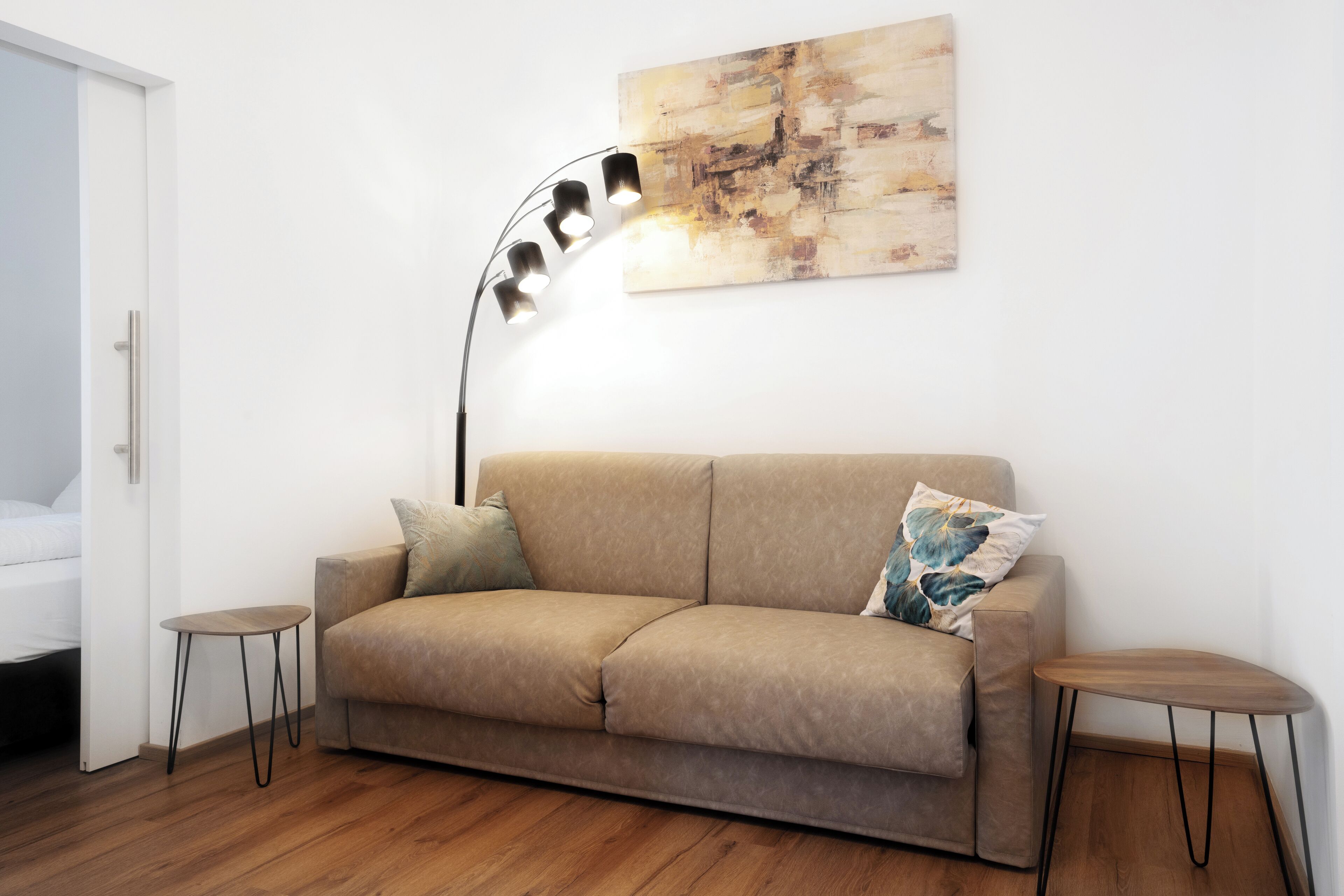 Ein modernes Wohnzimmer mit einem beigefarbenen Sofa, zwei kleinen Beistelltischen, einer Stehlampe und einem großen abstrakten Gemälde an der Wand.