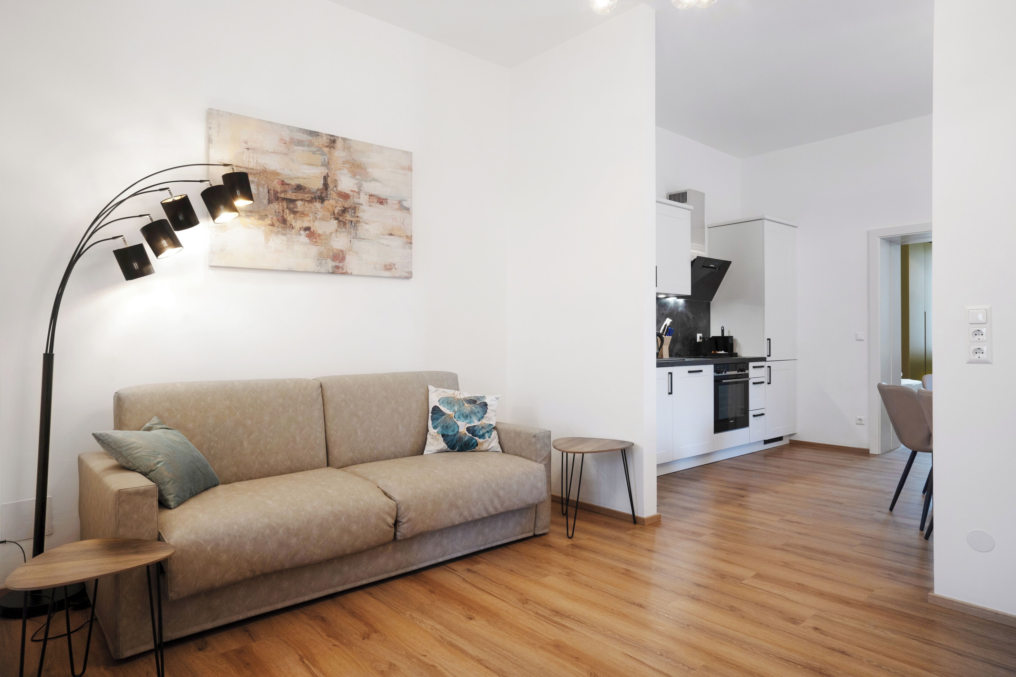 A modern living room with a sofa, a coffee table, and a floor lamp. There is a kitchen area with cabinets and a stove in the background.