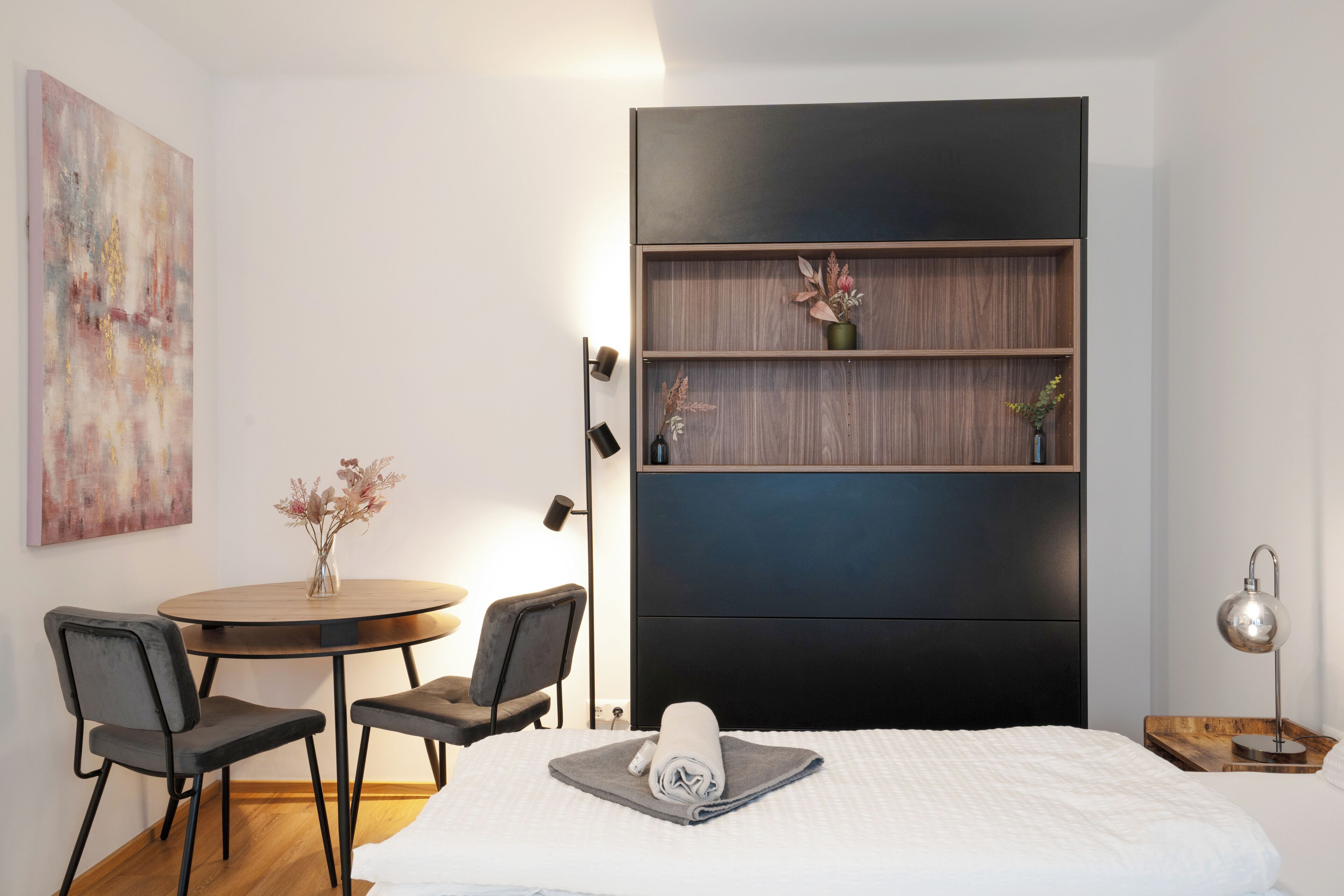 A modern hotel room with a bed, a round dining table with chairs, a small side table with a lamp, and a wall-mounted shelf with decorative items.