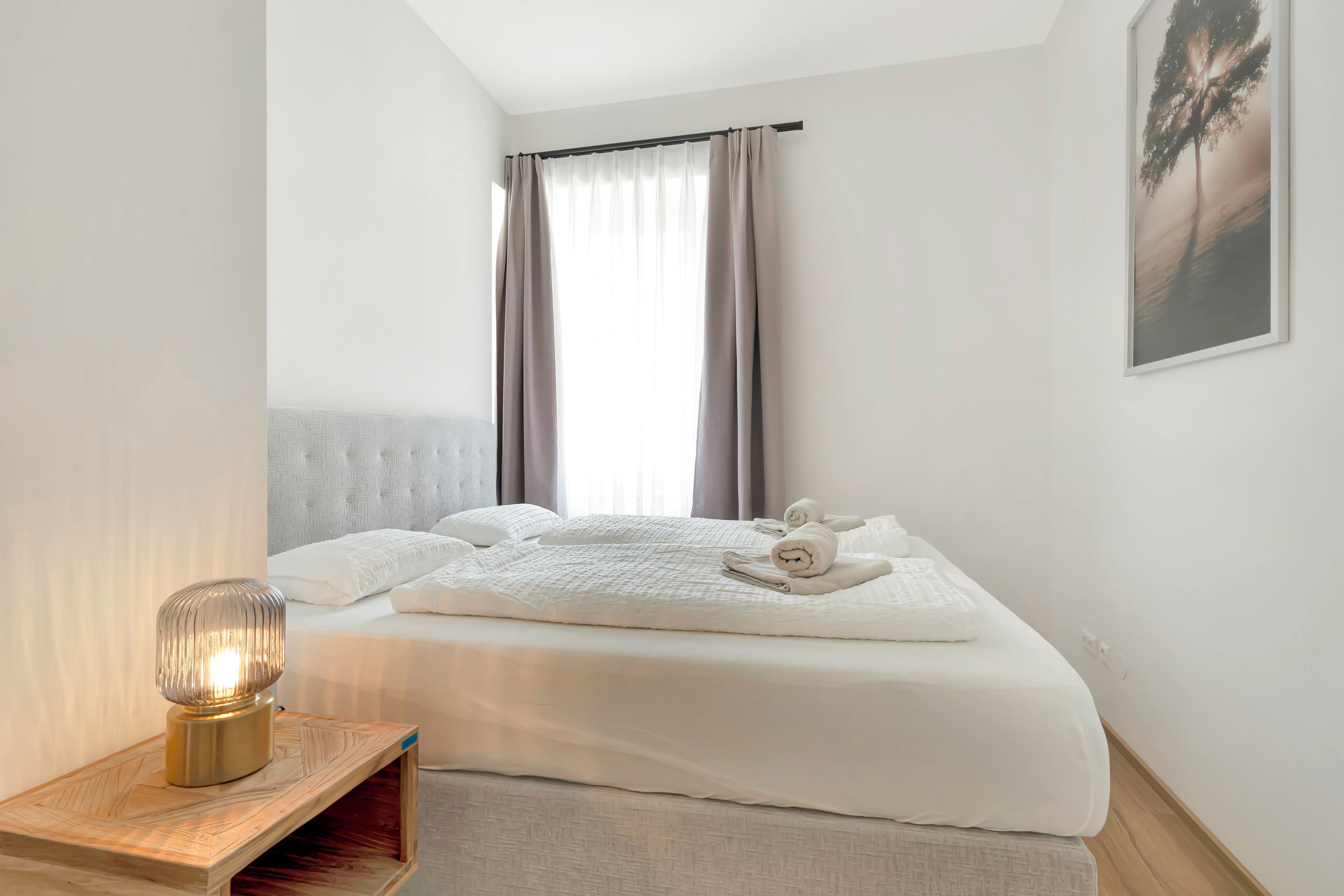 Ein ordentlich bezogenes Bett mit weißen Leinentüchern, ein Nachttisch mit Lampe und ein großes Fenster mit Vorhängen.