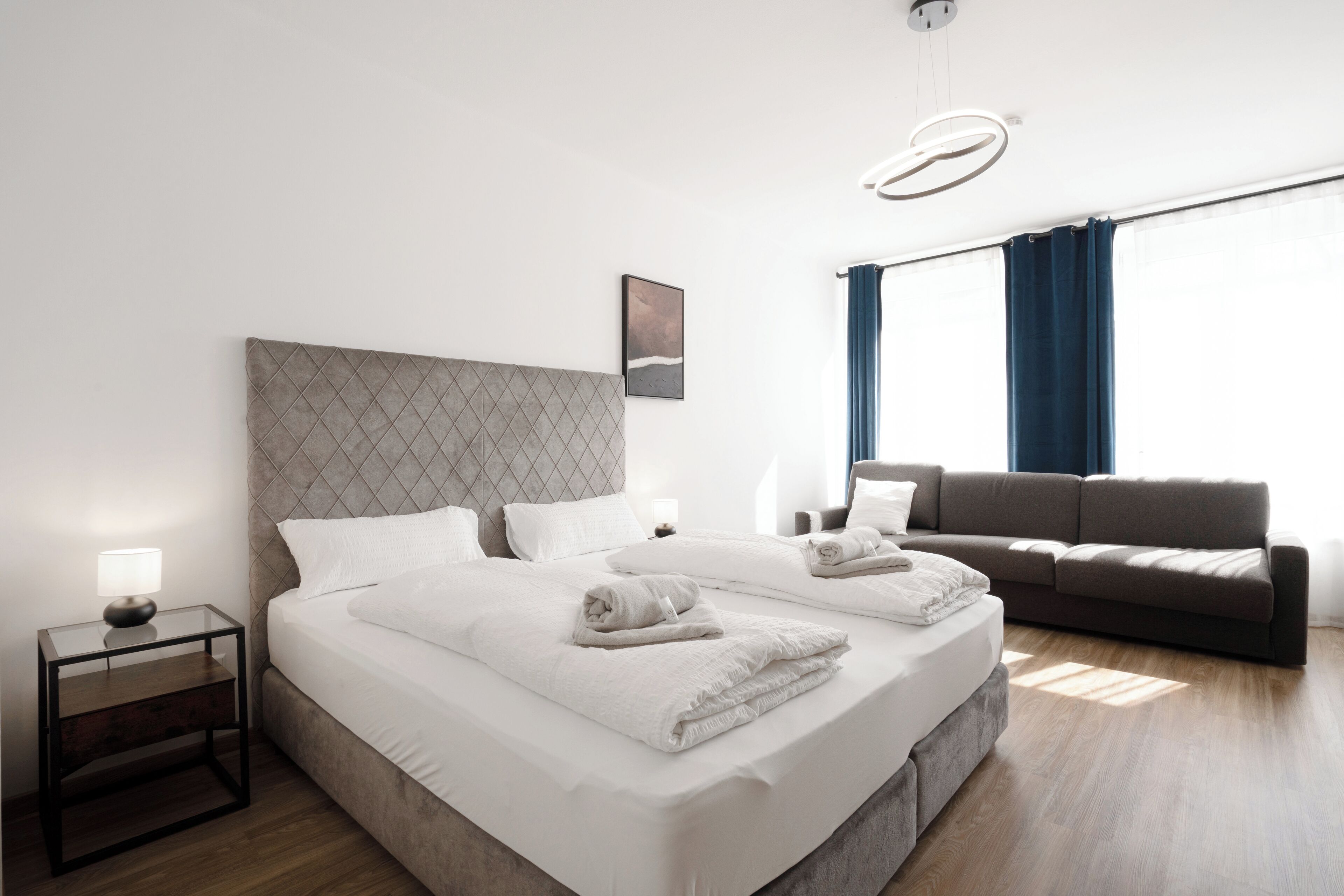 A modern hotel room with a large bed, a sofa, a nightstand, and a wall-mounted light fixture.