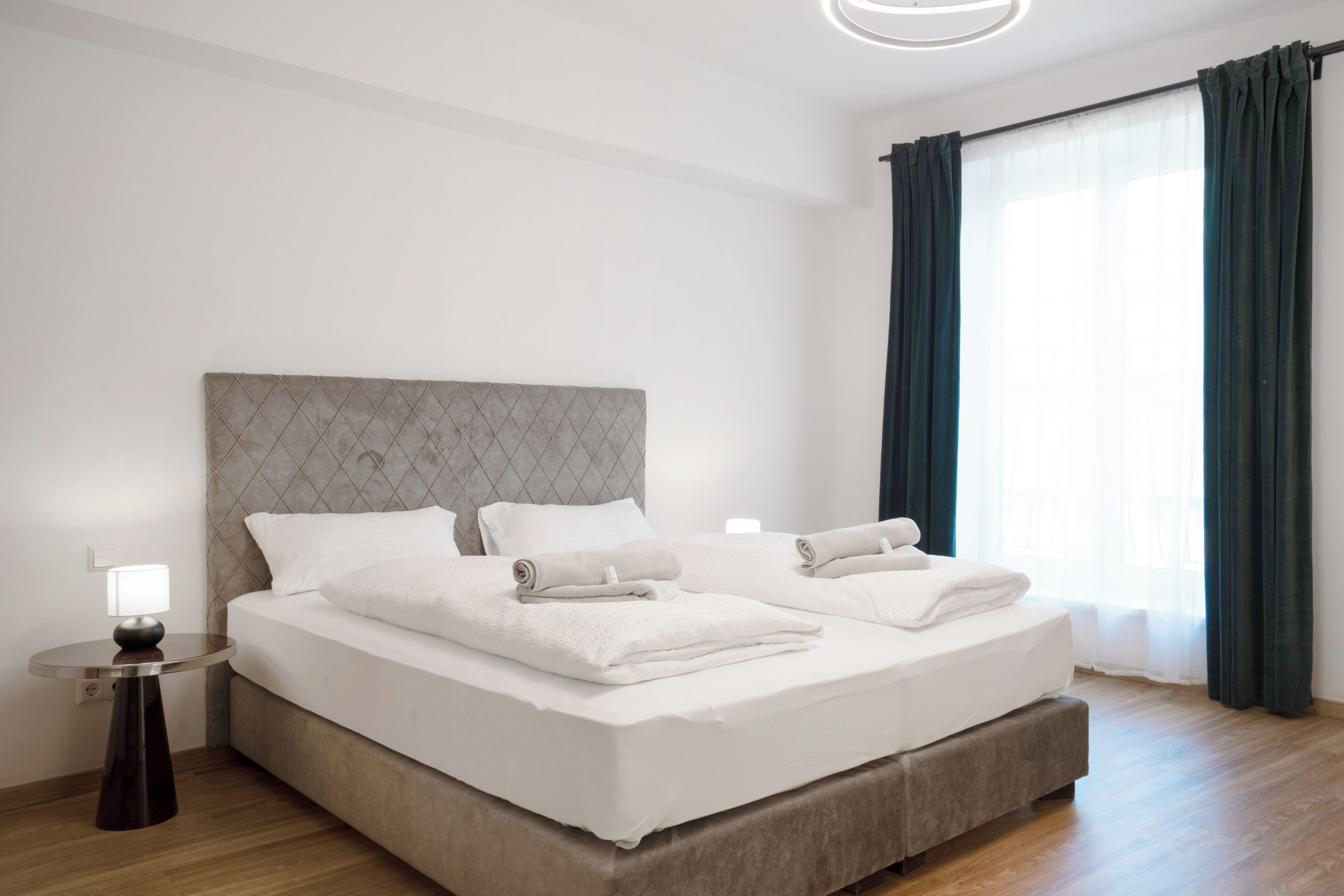 A modern hotel room with a large bed, a bedside table with a lamp, and a view of the outdoors through a window with curtains.