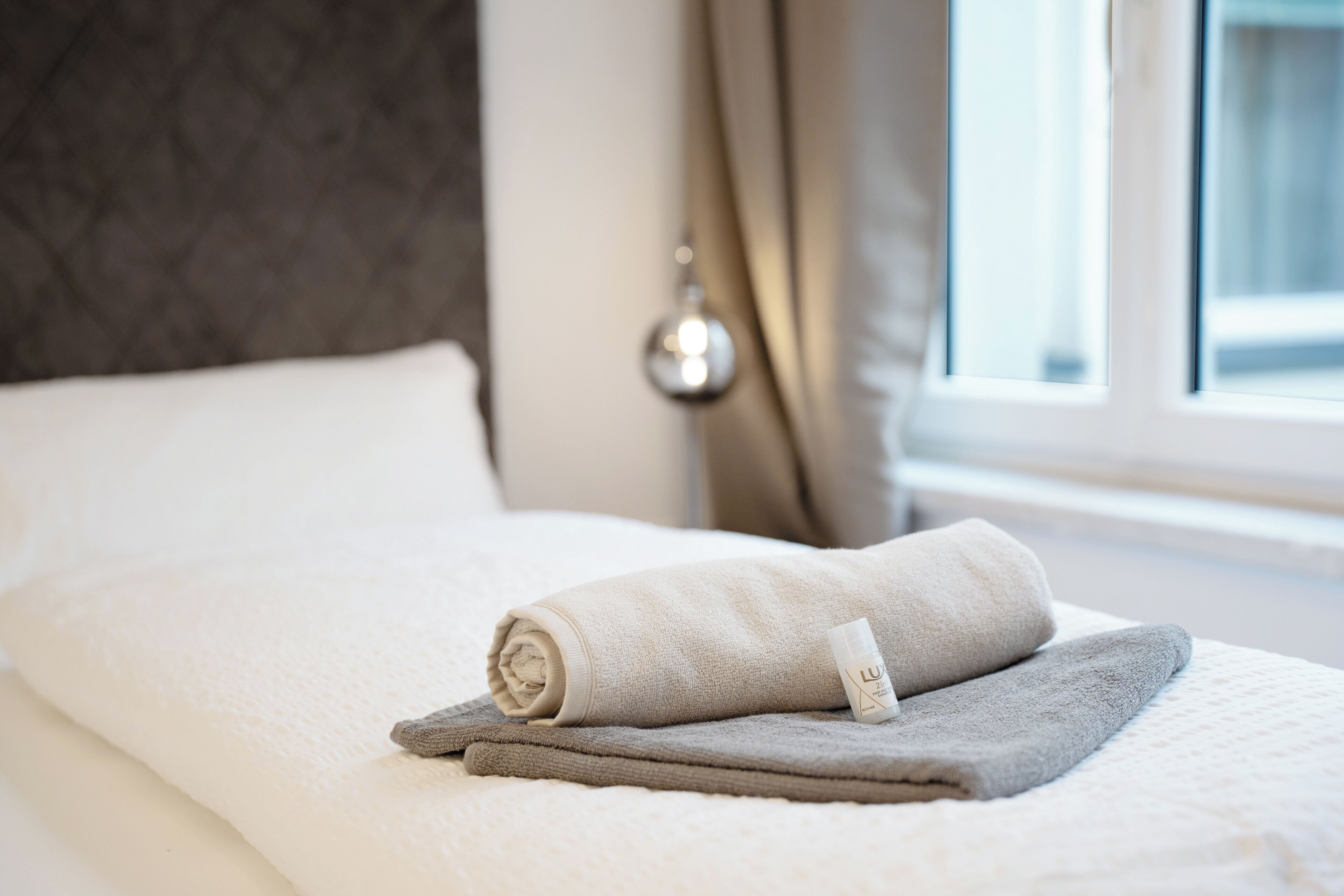 A neatly made bed with a folded towel and a bottle of lotion, a window with curtains, and a wall-mounted light fixture.