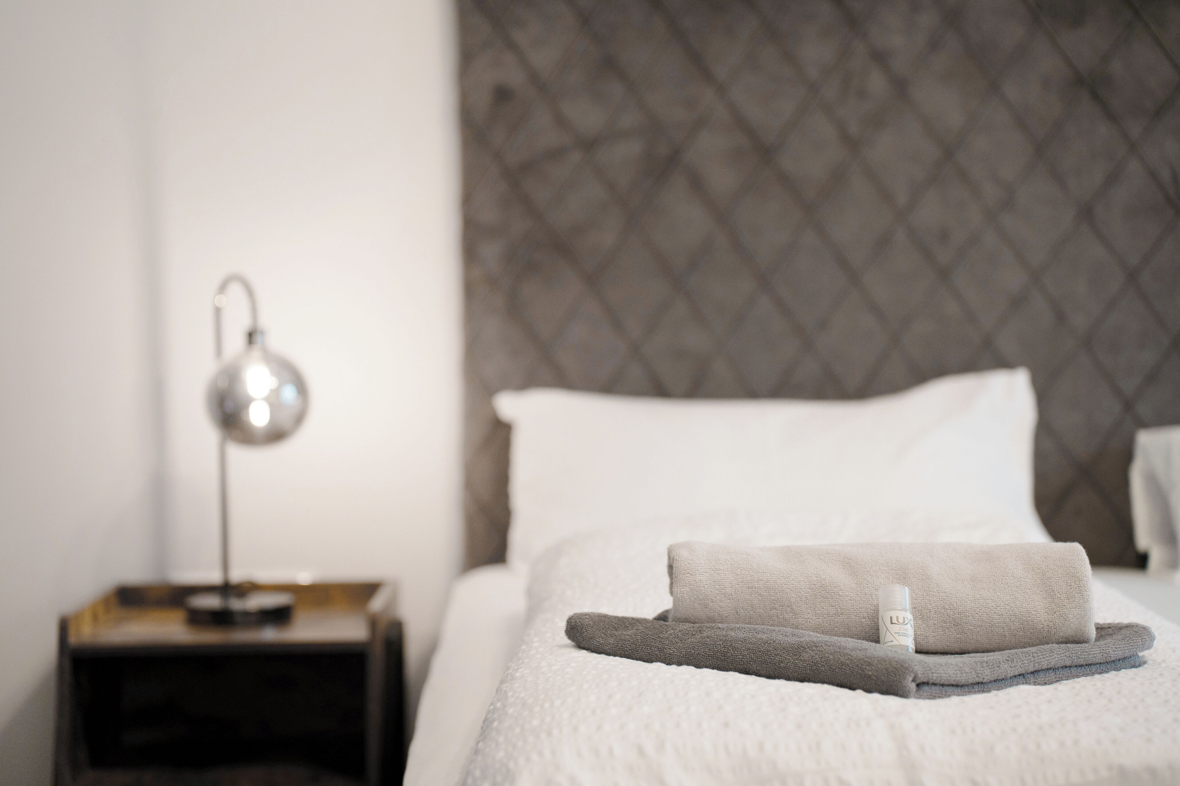 Una cama bien hecha con una toalla doblada y una botella de loción, una lámpara de noche y un cabecero con textura.
