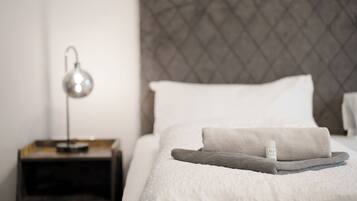 Una cama bien hecha con una toalla doblada y una botella de loción, una lámpara de noche y un cabecero con textura.