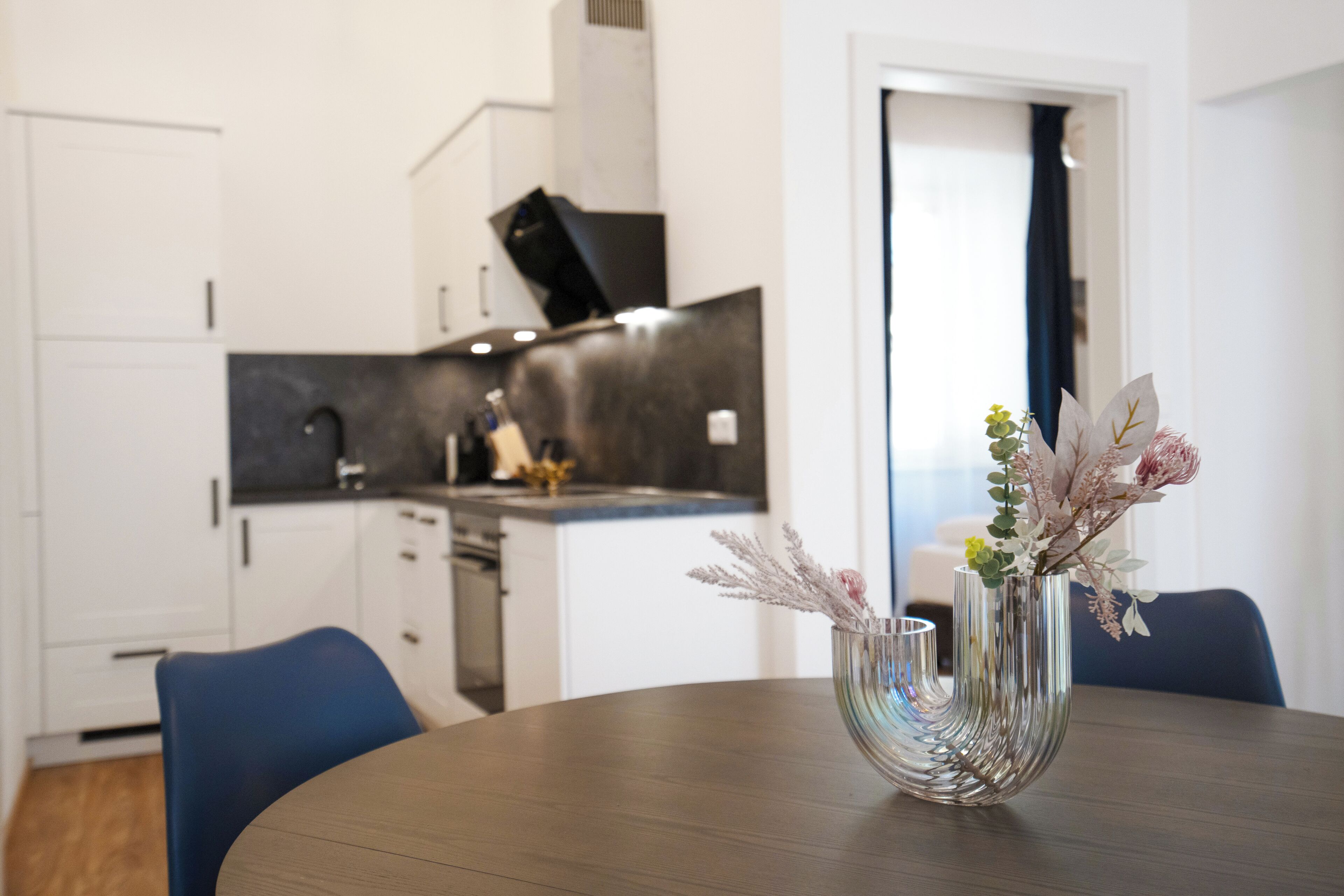 Et moderne køkken med hvide skabe og en mørk bordplade. En vase med blomster står på et træbord.