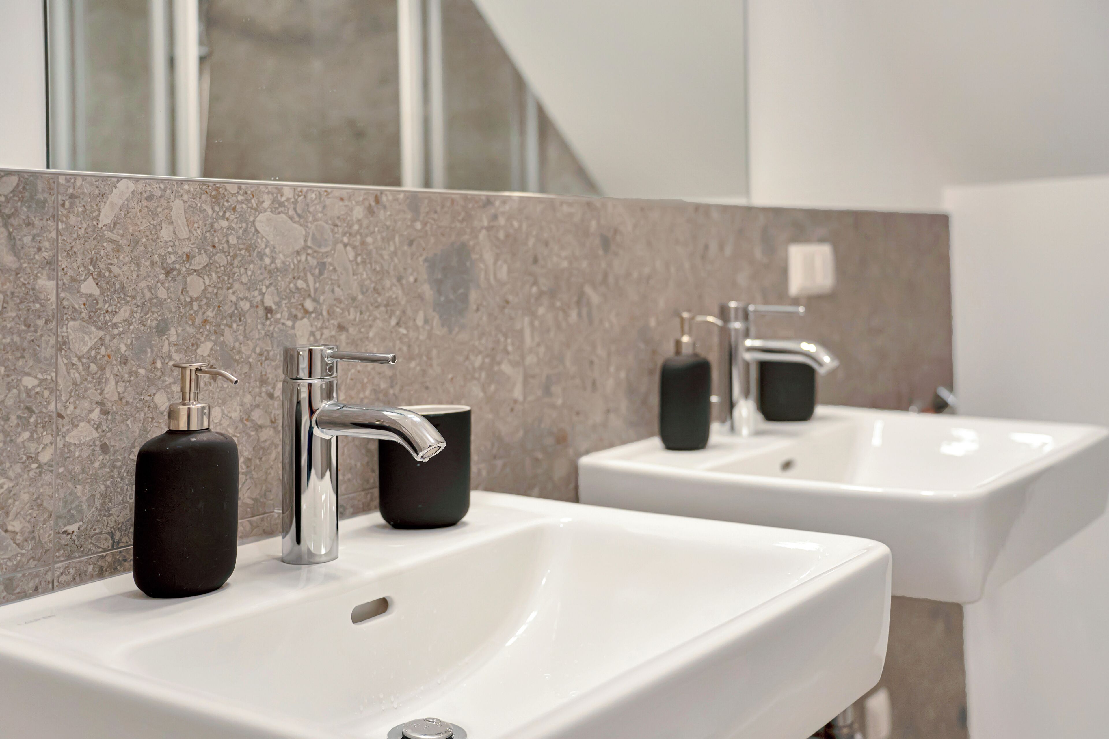 Ein modernes Badezimmer mit zwei weißen Waschbecken, schwarzen Seifenspendern und einer Marmor-Spritzschutzwand.