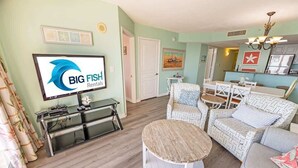 Un salon avec un téléviseur diffusant des publicités pour « BIG FISH Rentals », un coin repas avec une table et des chaises, et une cuisine équipée d’un réfrigérateur et de placards.