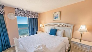 Un lit bien fait, avec du linge de lit blanc et une serviette pliée, une table de chevet blanche avec une lampe, et une vue sur la mer par la fenêtre.