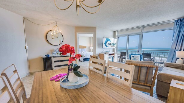 A modern living room with a wooden floor, a dining table with chairs, a chandelier, a TV, and a large window with a view of the sea. - Amazing Baywatch 3BR with Pools Direct Ocean Front (North Myrtle Beach)