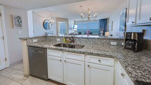 A kitchen with granite countertops, a dishwasher, a coffee maker, and a view of the ocean through large windows. - Amazing Baywatch 3BR with Pools Direct Ocean Front (North Myrtle Beach)
