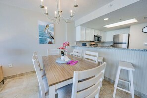 A dining area with a wooden table, white chairs, a chandelier, a kitchen with white cabinets, a refrigerator, and a microwave.