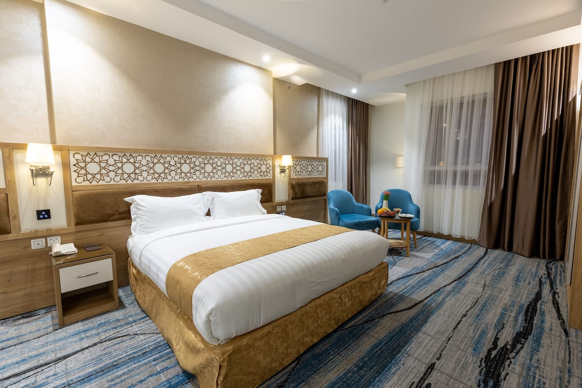 A hotel room with a large bed, a nightstand, a blue armchair, a small table with a fruit bowl, and a large window with curtains.