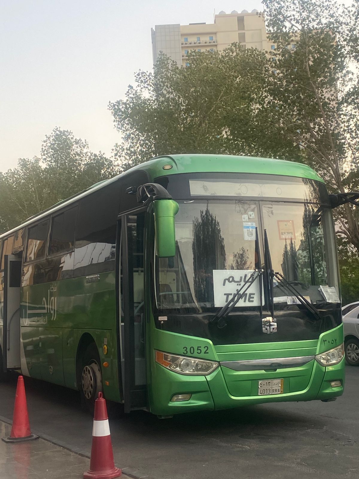 Un autobús verde con el número 3052 estacionado en una calle, con conos de tráfico y árboles al fondo.