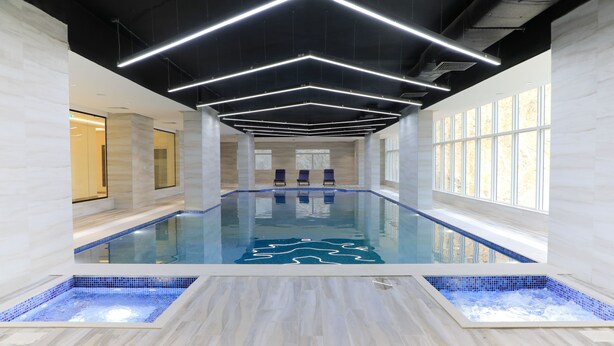 En moderne indendørs swimmingpool med geometriske loftslamper, glasvægge og flisegulv.